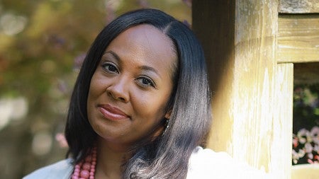 Kimberly Johnson smiling, wearing a white shirt, light beige cardigan and pink necklace while leaning up against a wooden post.